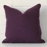 A square cushion in a delightful plum tone rests against a white wall. The linen material appears to be of exceptional quality.