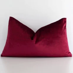 Burgundy solid colour cushion leans elegantly against a white wall. It has been crafted from a high quality velvet material and has a rectangle shape.