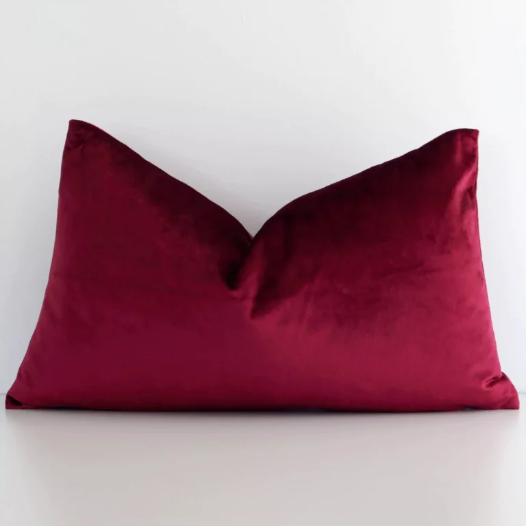 Burgundy solid colour cushion leans elegantly against a white wall. It has been crafted from a high quality velvet material and has a rectangle shape.