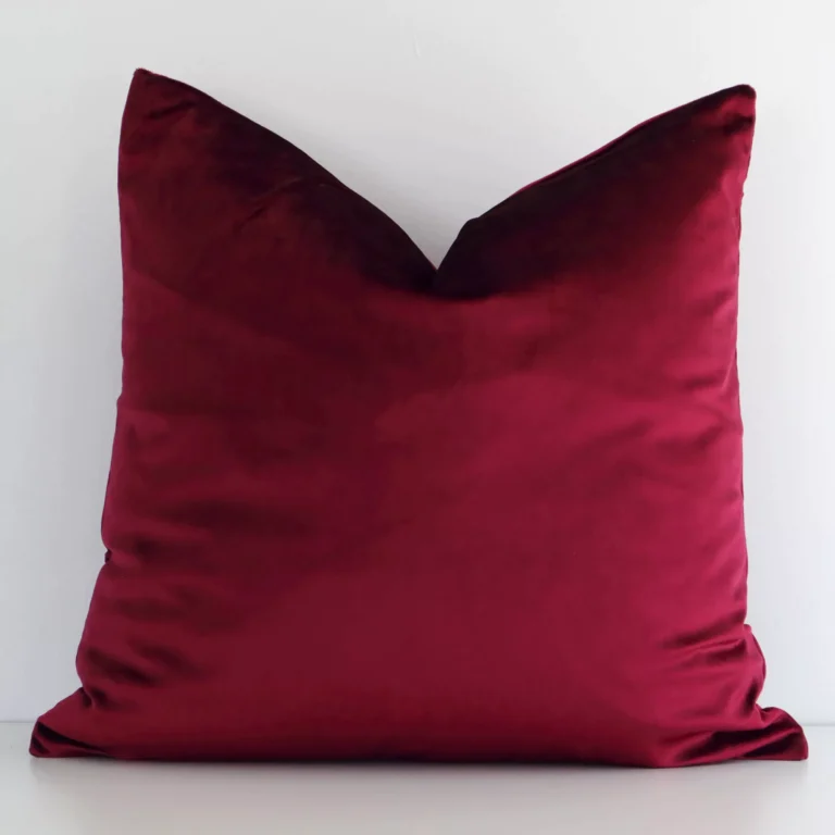A white wall that has a solid coloured burgundy cushion cover positioned in front of it. It has an exquisite velvet material and a lovely square shape.