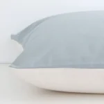 A solid-coloured soft teal cushion cover is laid flat. This angle shows the side of the linen fabric and its large size.