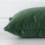 A brick wall that has a dark sage cushion cover positioned in front of it. It has an exquisite velvet material and a lovely rectangle shape.