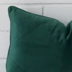 Enlarged shot of the corner of this rectangle cushion cover in emerald green colour is shown against a brick wall. The image shows the quality and craftsmanship of the velvet material.
