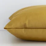 The edge of this velvet square cushion in gold mustard is shown. The shot shows the solid-coloured design and the front and rear panels.
