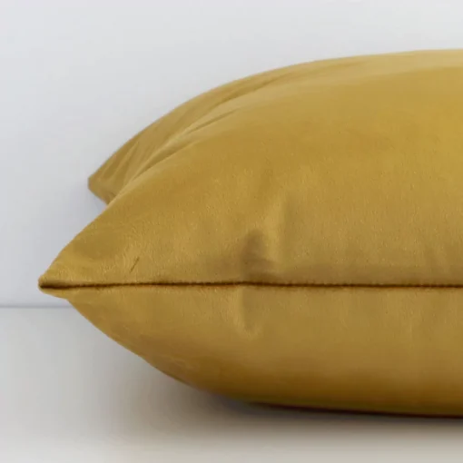 The edge of this velvet square cushion in gold mustard is shown. The shot shows the solid-coloured design and the front and rear panels.