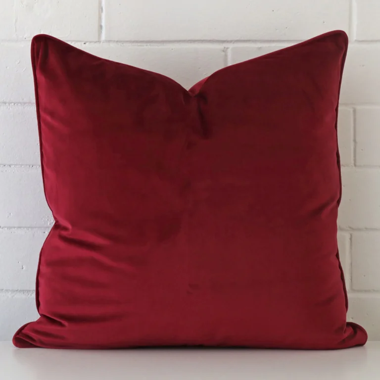 A large cushion in a delightful maroon tone rests against a white wall. The velvet material appears to be of exceptional quality.