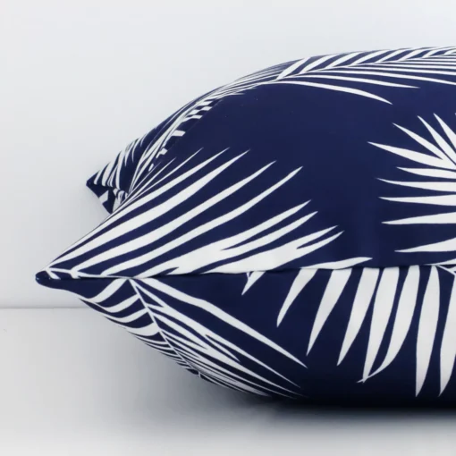 A palm printed navy cushion cover is laid flat. This angle shows the side of the outdoor fabric and its square shape.