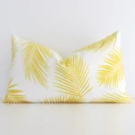 Yellow palm printed cushion leans elegantly against a white wall. It has been crafted from a high quality outdoor material and has a rectangle shape.