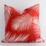 Red palm printed cushion leans elegantly against a white wall. It has been crafted from a high quality outdoor material and has a square shape.