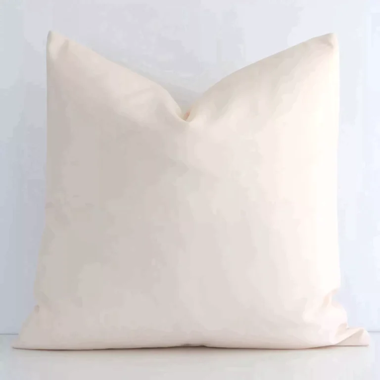 Solid-coloured square beige cushion positioned in front of a white wall. It is made of outdoor fabric.