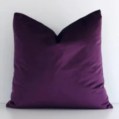 White wall with a solid-coloured plum cushion laying against it. It has a distinctive velvet fabric and has a square shape.