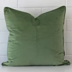 Sage cushion positioned in front of a brick wall. It has large dimensions and is made from a velvet material.