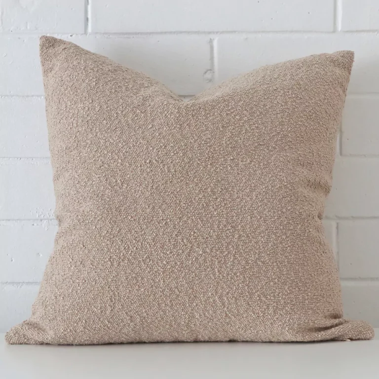 A pretty boucle cushion cover is shown against a brick wall. It features a large shape and a beige colour finish.